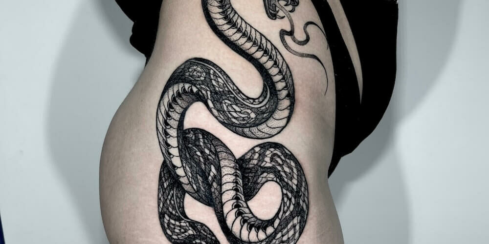 Foto de una mujer joven con el lateral del cuerpo (muslo hasta cadera) tatuado con estilo de tatuaje blackwork de una serpiente por Carlos cuervo, tatuador de kaifa´s tattoo studio en Madrid
