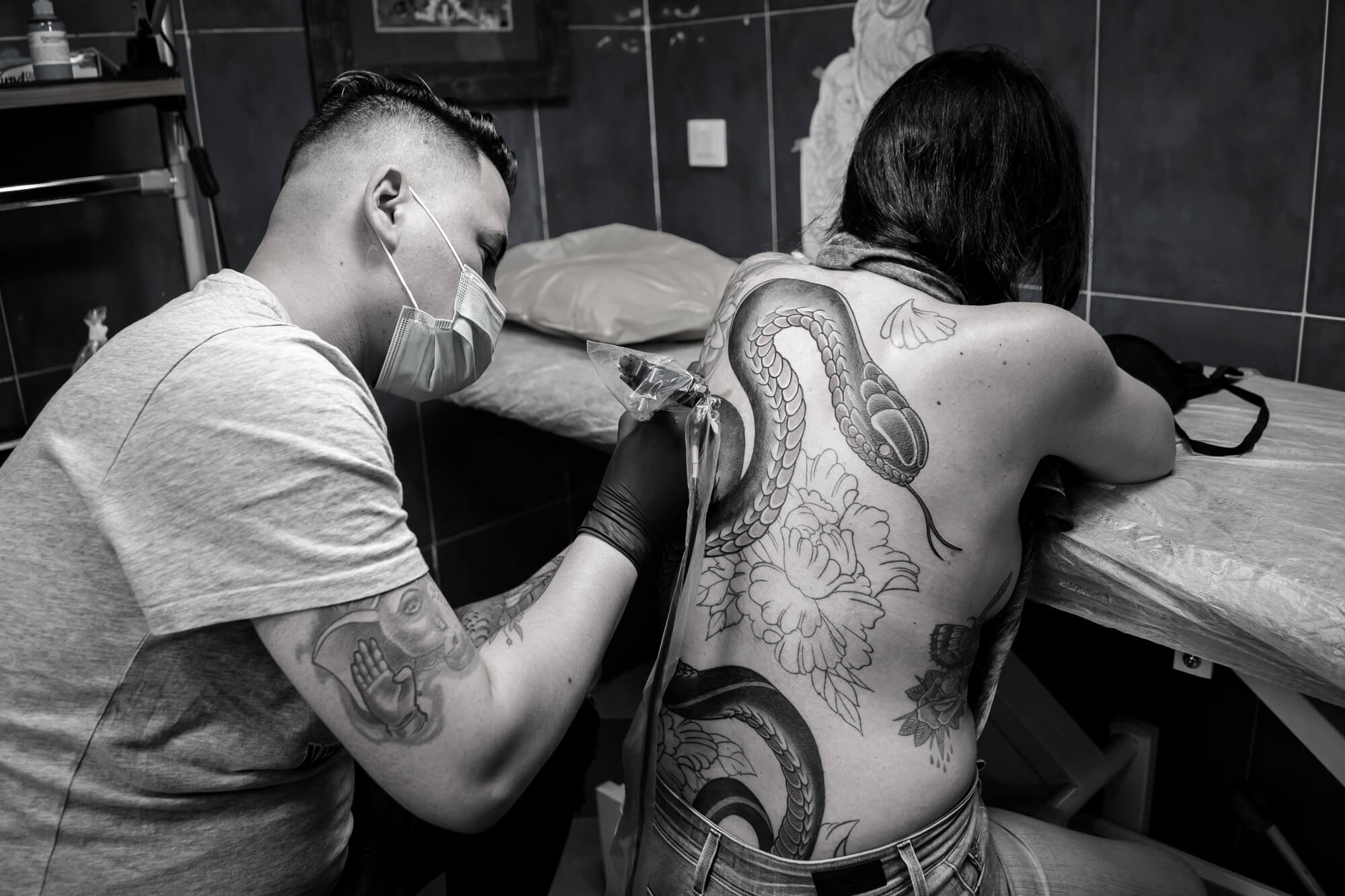 foto en blanco y negro en el Estudio de tatuajes y piercings Kaifa´s Tattoo Madrid en la zona de Chamberí. Se ve a un tatuador con mascarilla tatuando la espalda de un cliente con tinta vegana